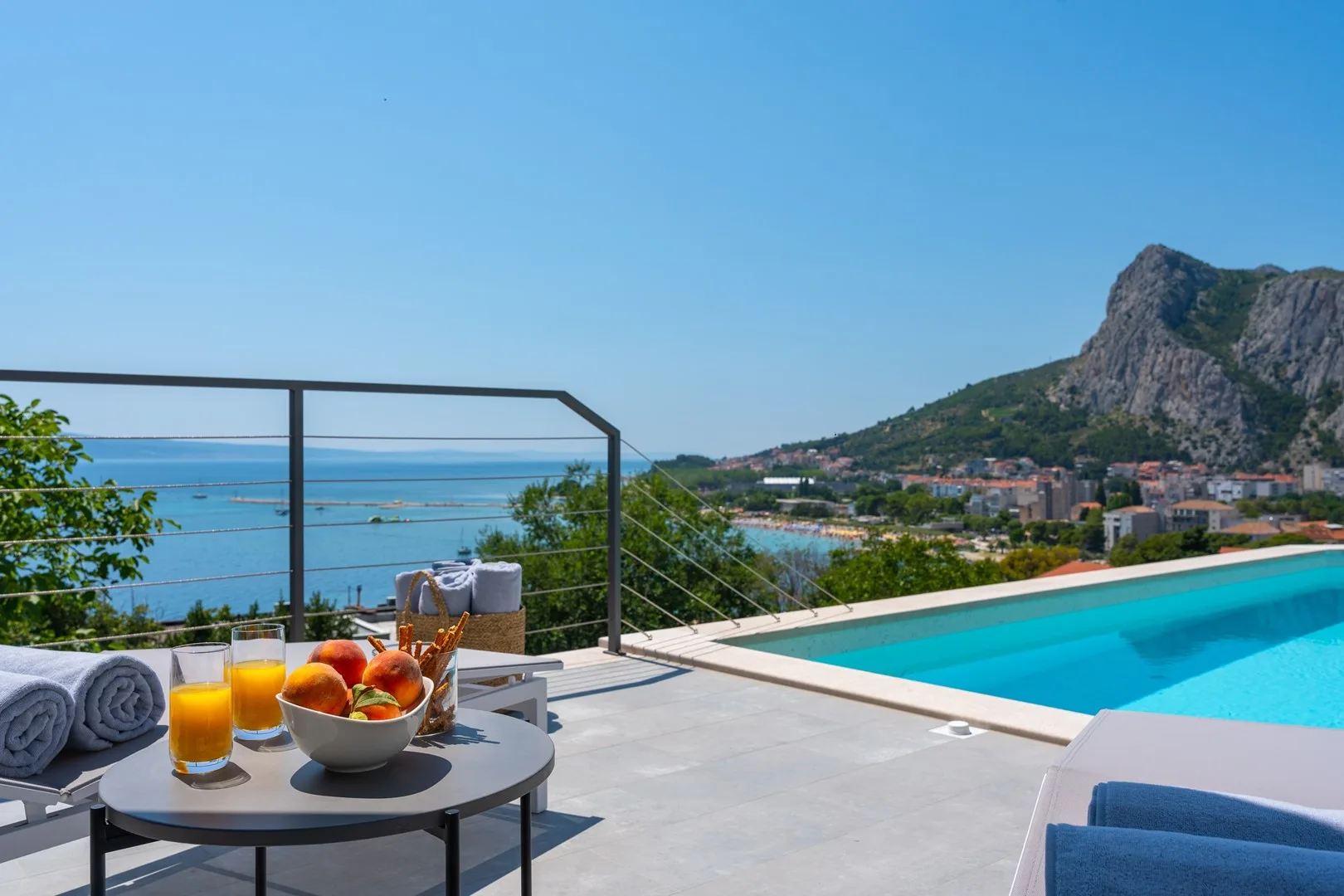 A poolside terrace overlooks the sea, with outdoor seating and a fruit bowl.