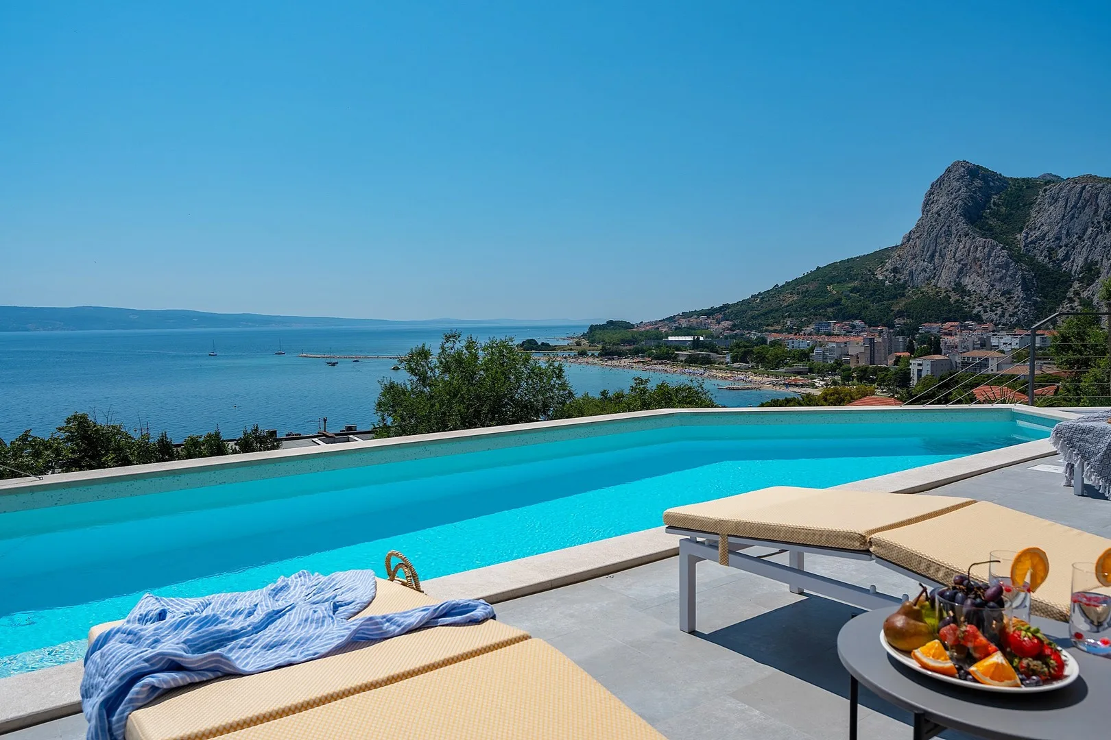 Poolside sun deck with loungers overlooking the sea and a coastal town.