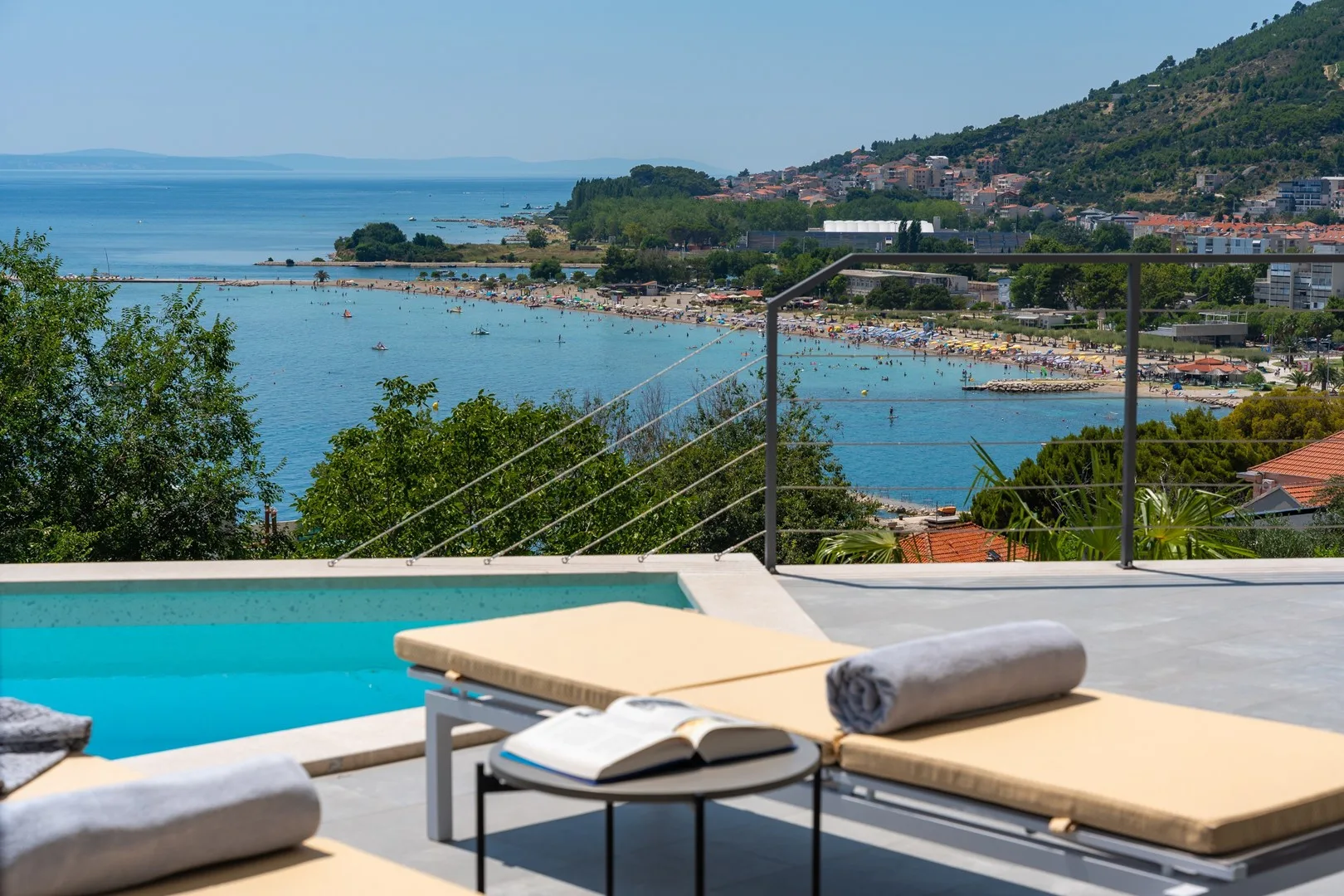 A poolside sun deck overlooks the sea, beach, and hillside town.