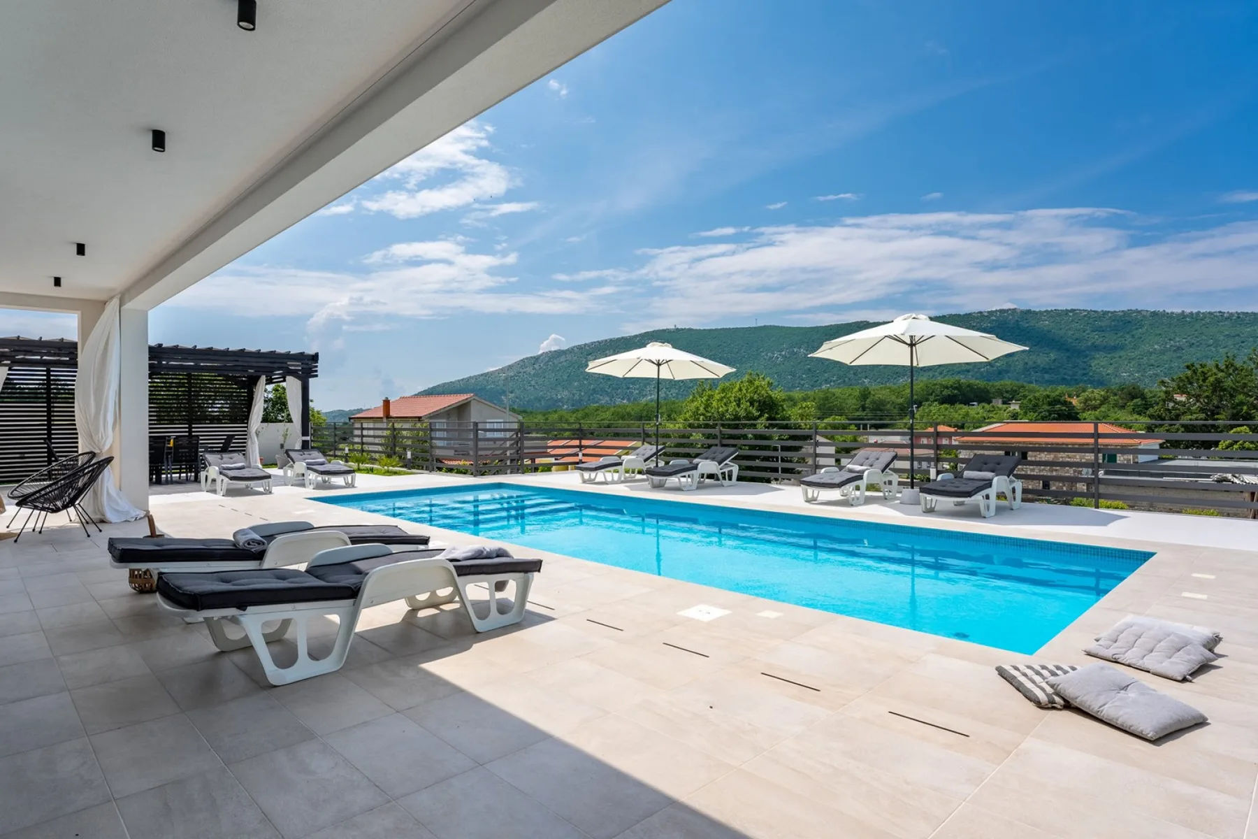 A swimming pool sits beside a tiled patio with loungers and umbrellas.
