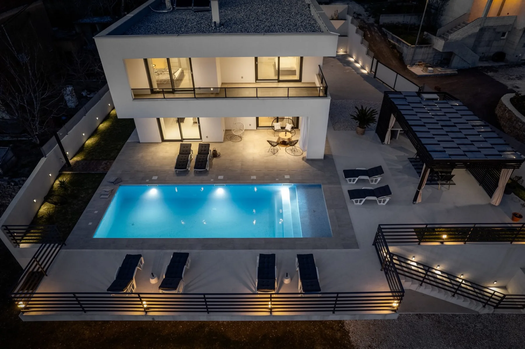 An illuminated outdoor pool and patio sit beside a modern house at night.