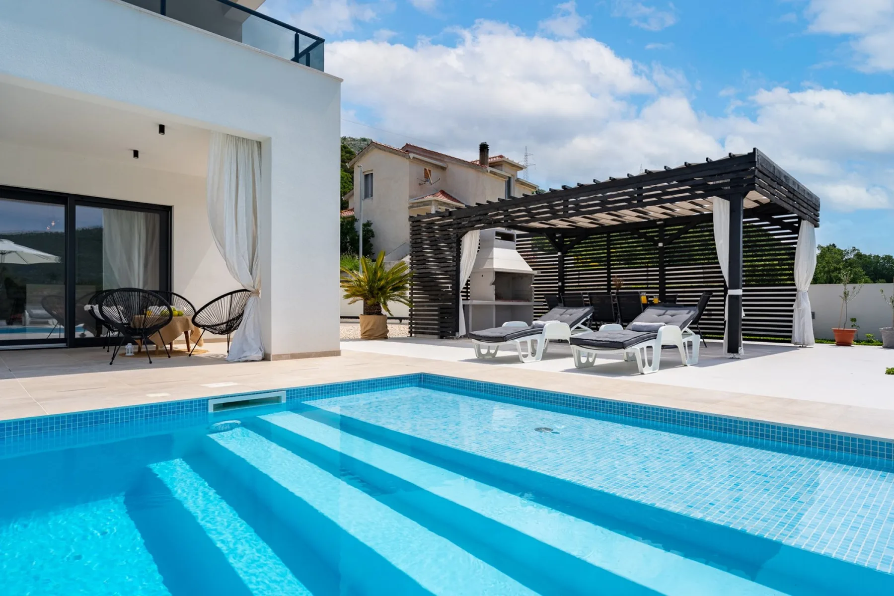 Una piscina exterior moderna junto a un edificio blanco y una zona de descanso con sombra.