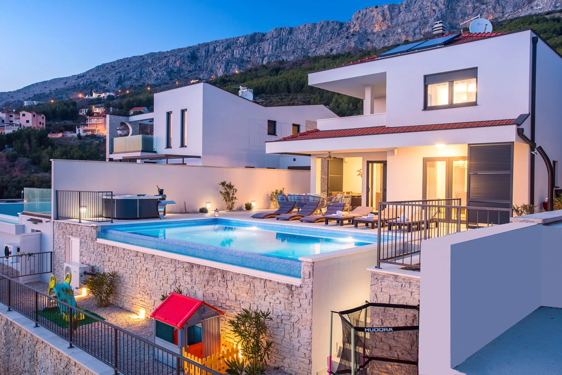 Modern villa buildings overlook a lit swimming pool and outdoor seating at dusk.