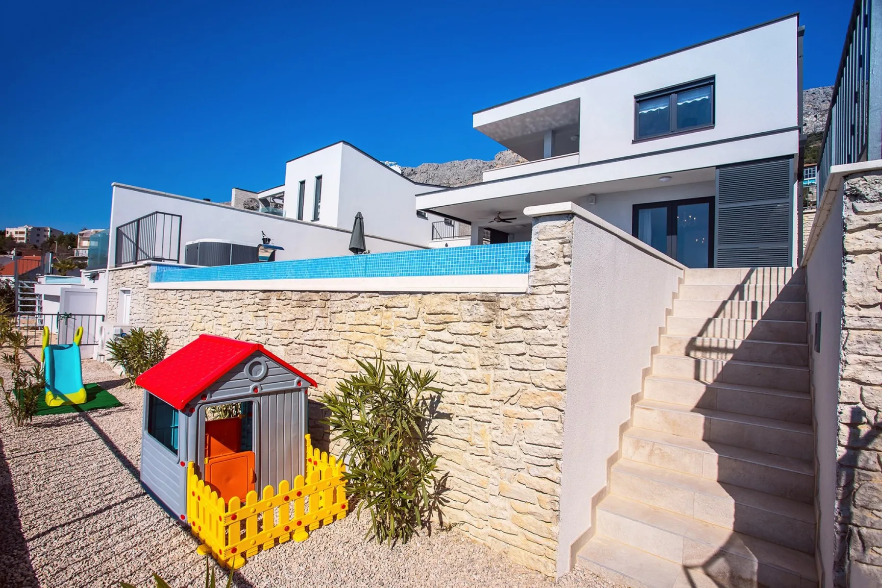 Outdoor children’s play area with a small playhouse and slide beside a stone wall.