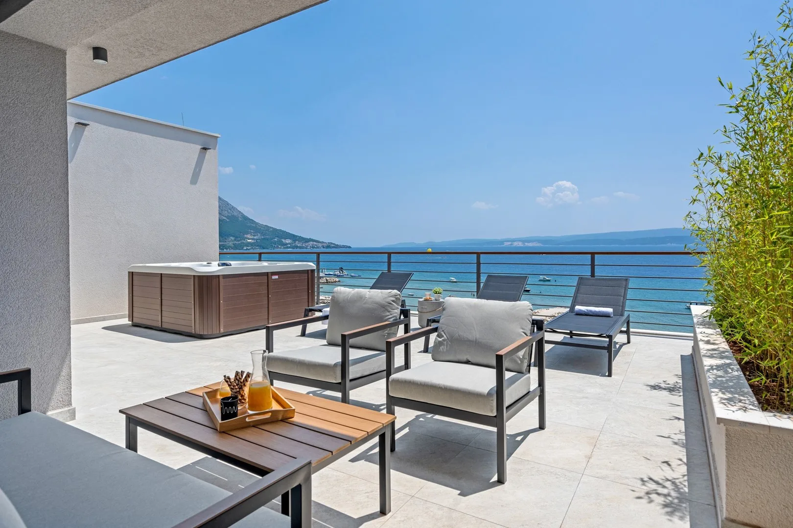 A sunlit terrace with lounge seating, a hot tub, and views of the sea.