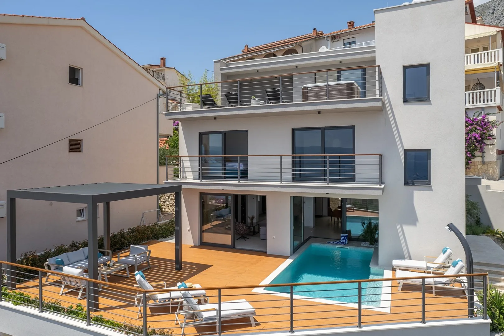 A modern multi-story villa overlooks a pool and a furnished outdoor deck.