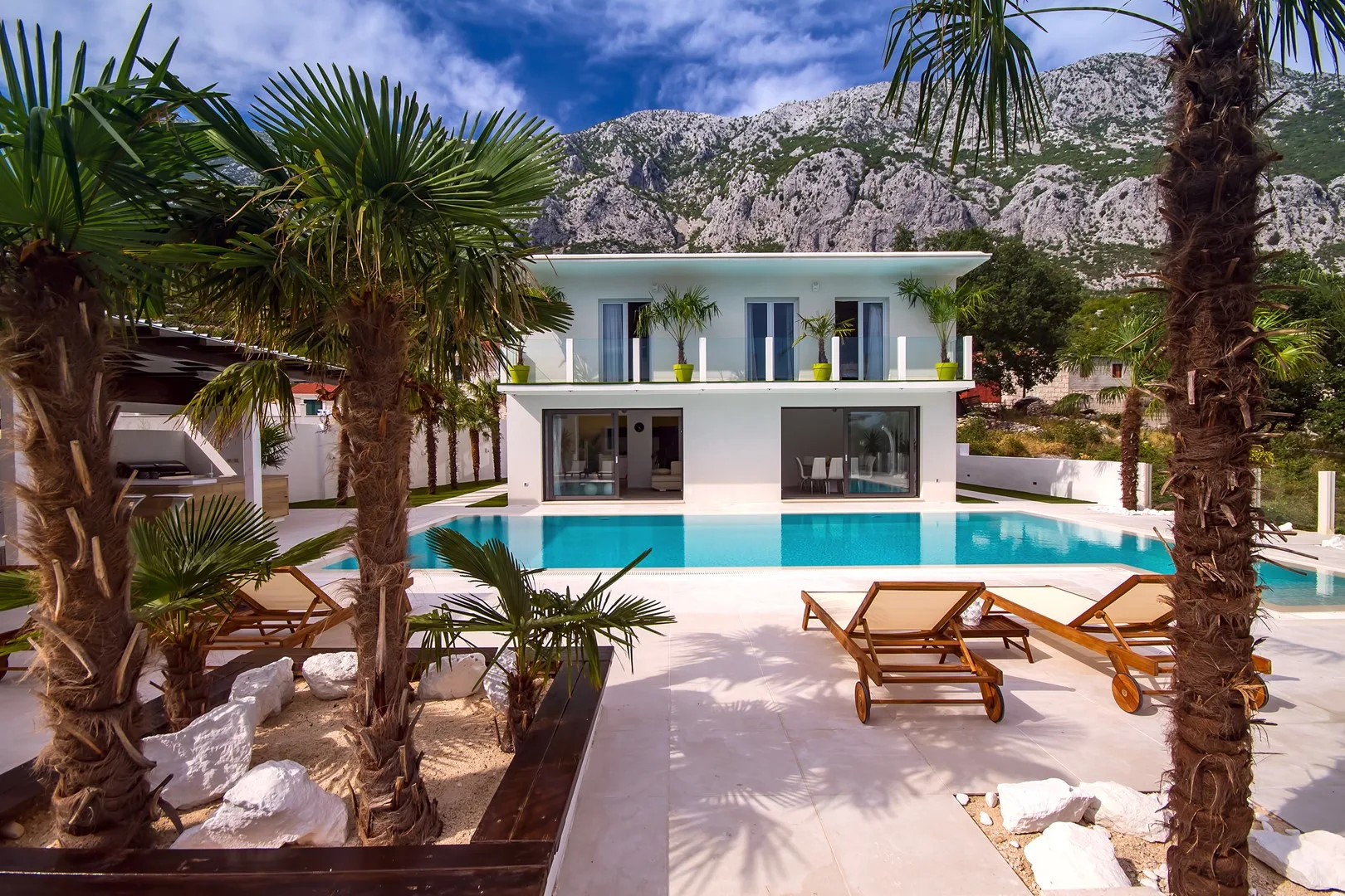 A modern house overlooks a swimming pool, with mountains visible behind it.