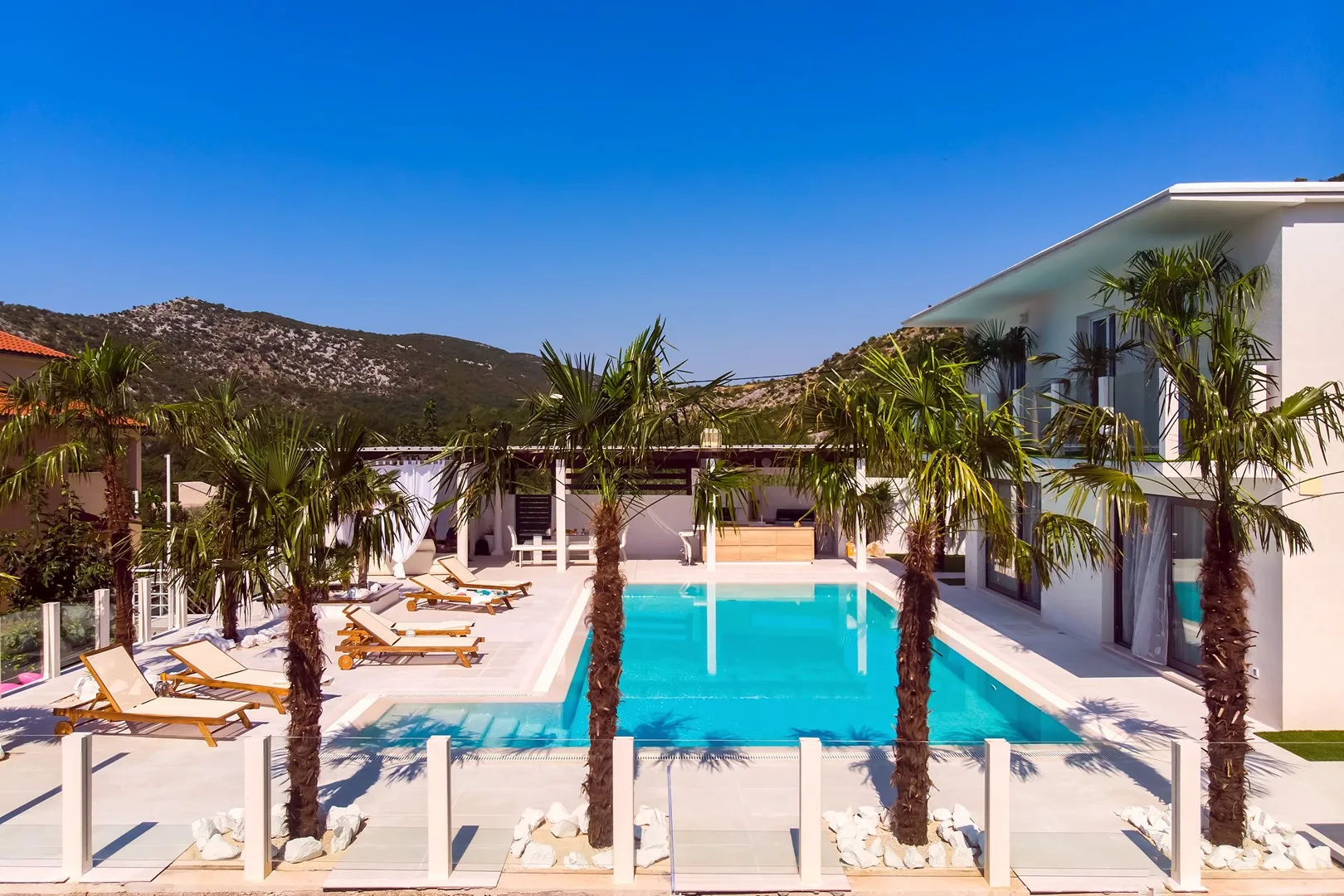 An outdoor swimming pool beside a modern building, with palm trees and lounge seating.