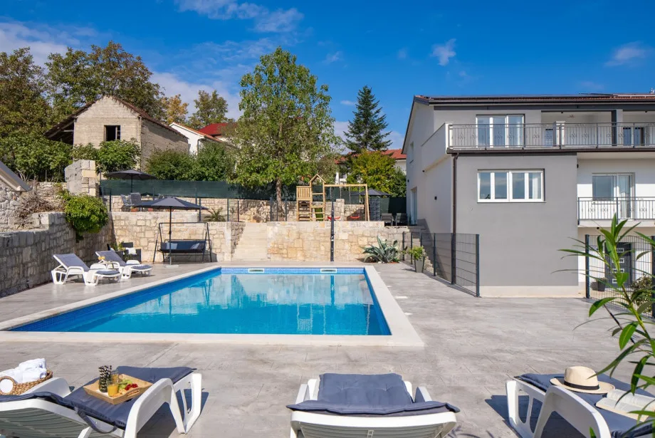 La zona de la piscina privada de 50 m² con tumbonas, la casa principal y la zona de juegos situada en una parte elevada.