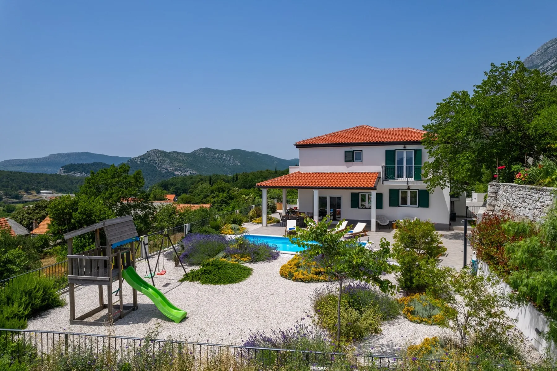 Una villa con piscina y zona de juegos está rodeada de jardines.