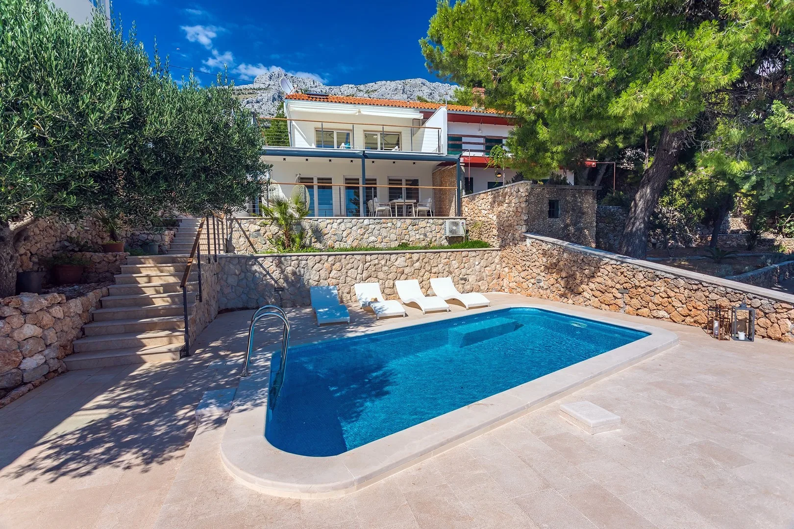 A swimming pool and sun loungers sit in front of a multi-level house.