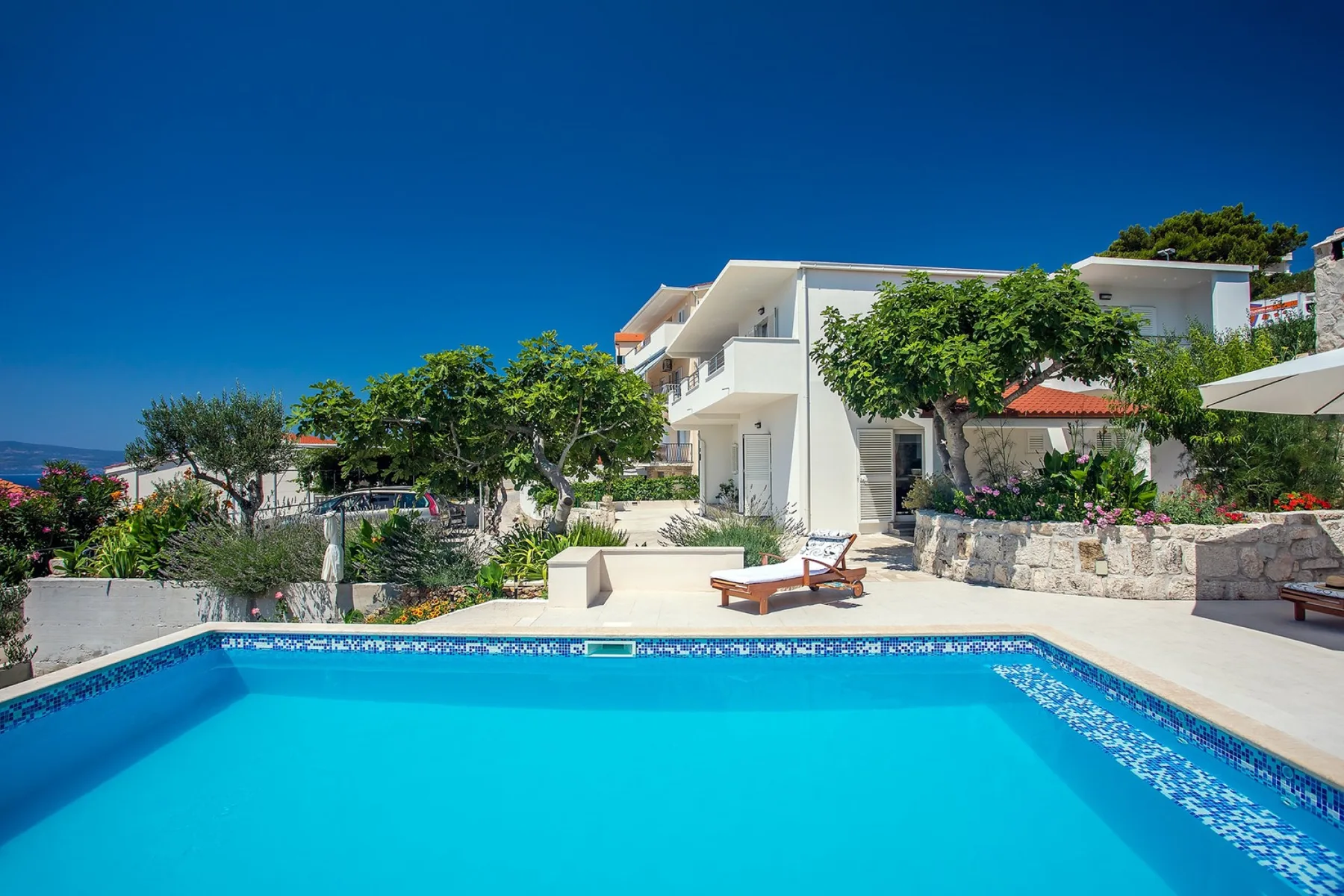A swimming pool fills the foreground of a sunny outdoor scene with seating and greenery.