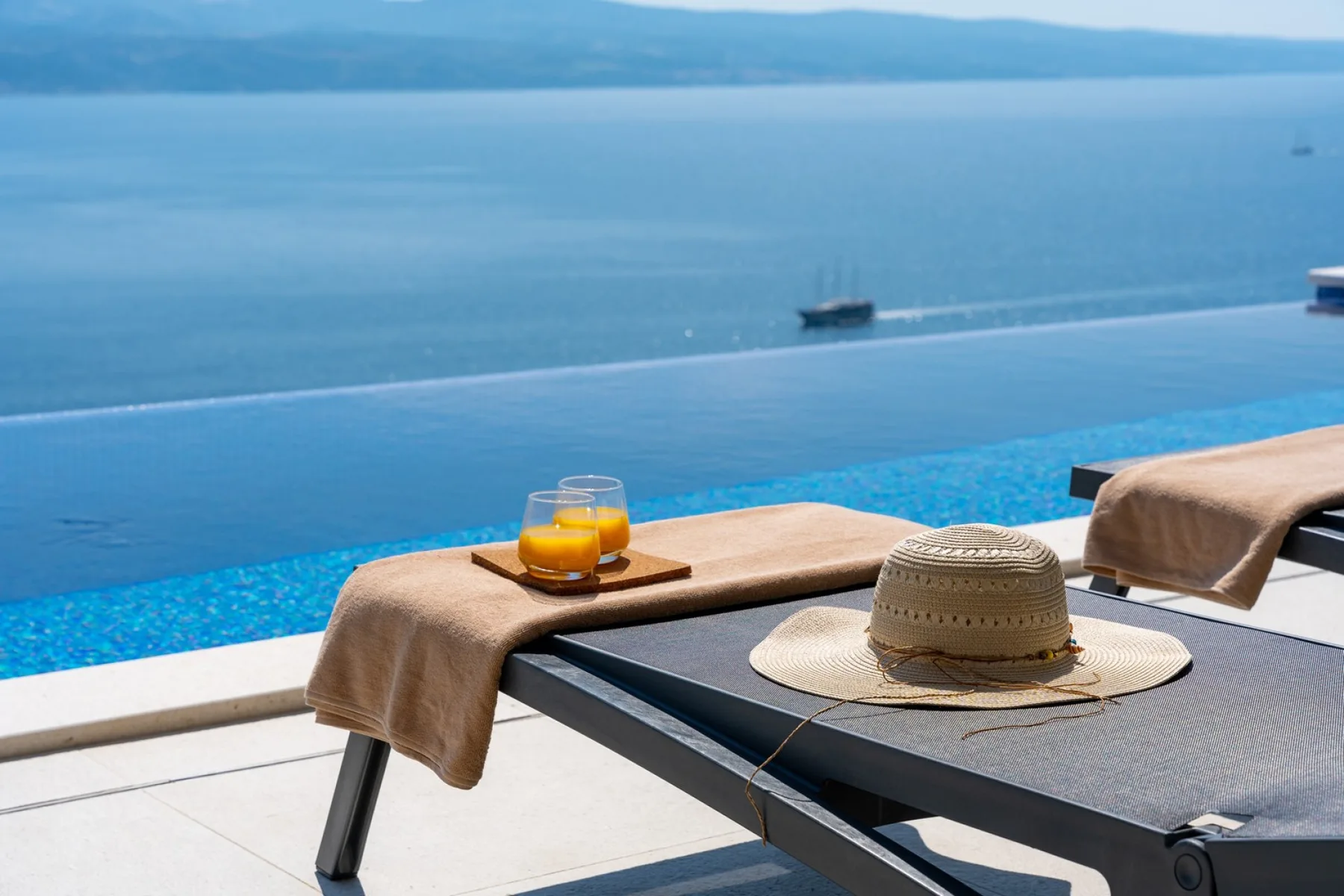 Una zona de descanso junto a la piscina mira al mar, con un sombrero y bebidas sobre una mesa.