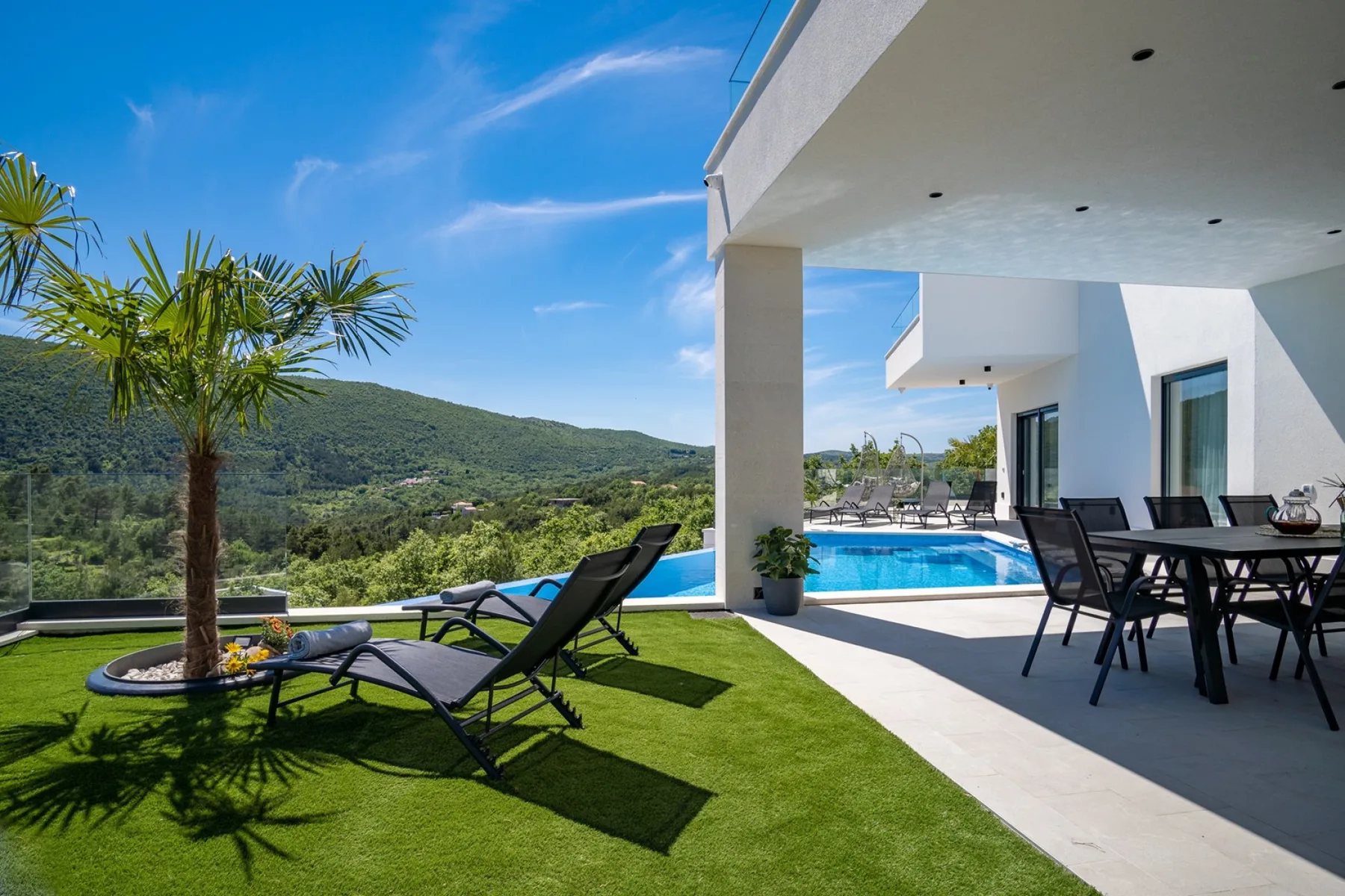 A sunny outdoor patio has lounge seating beside a swimming pool and green lawn.