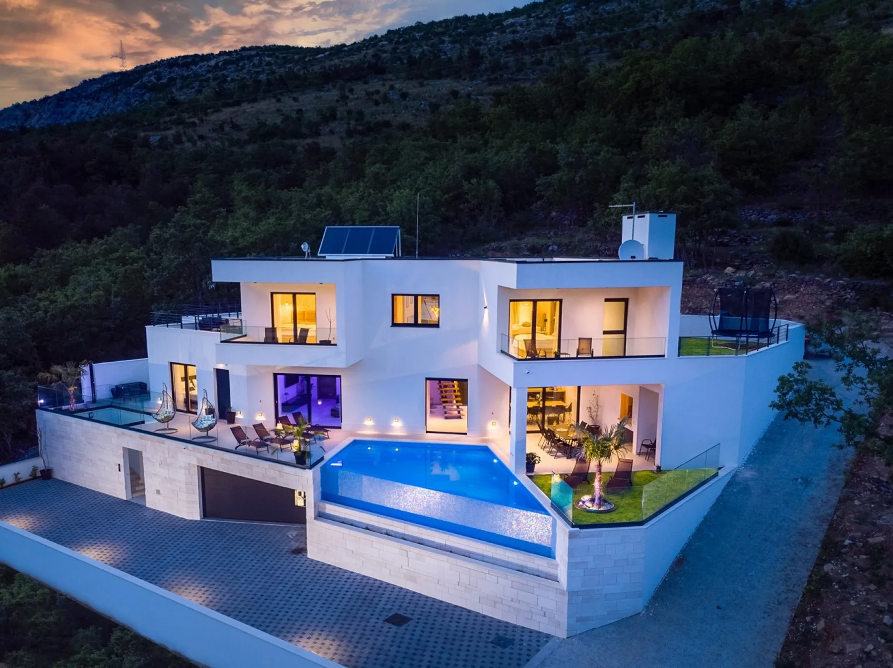 A modern white house with a swimming pool and paved outdoor area sits against a wooded hillside.