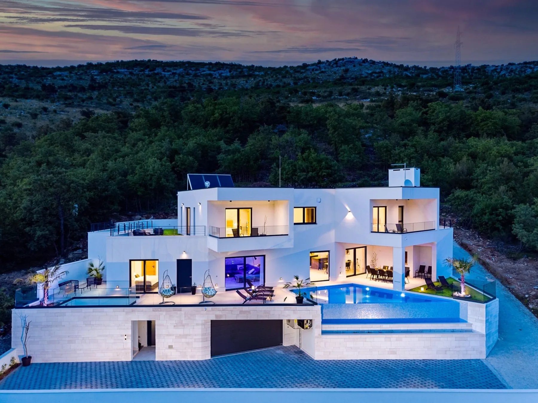 A modern villa sits beside a swimming pool, with wooded hills in the background.