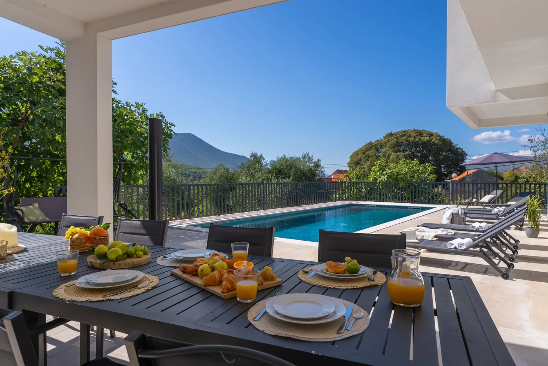 An outdoor dining table overlooks a swimming pool, trees, and a mountain.