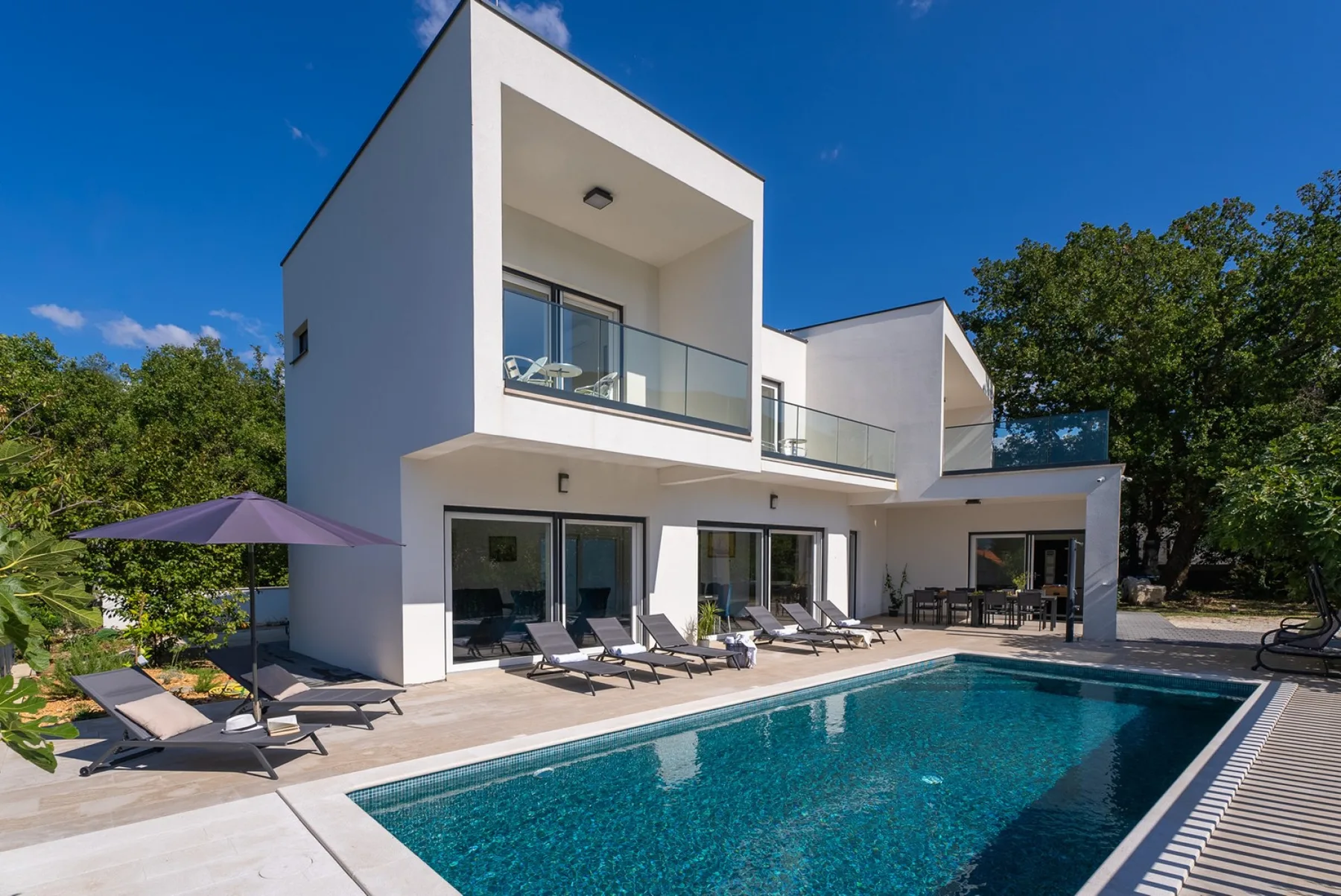 A modern villa opens onto a swimming pool, with outdoor seating on the patio.