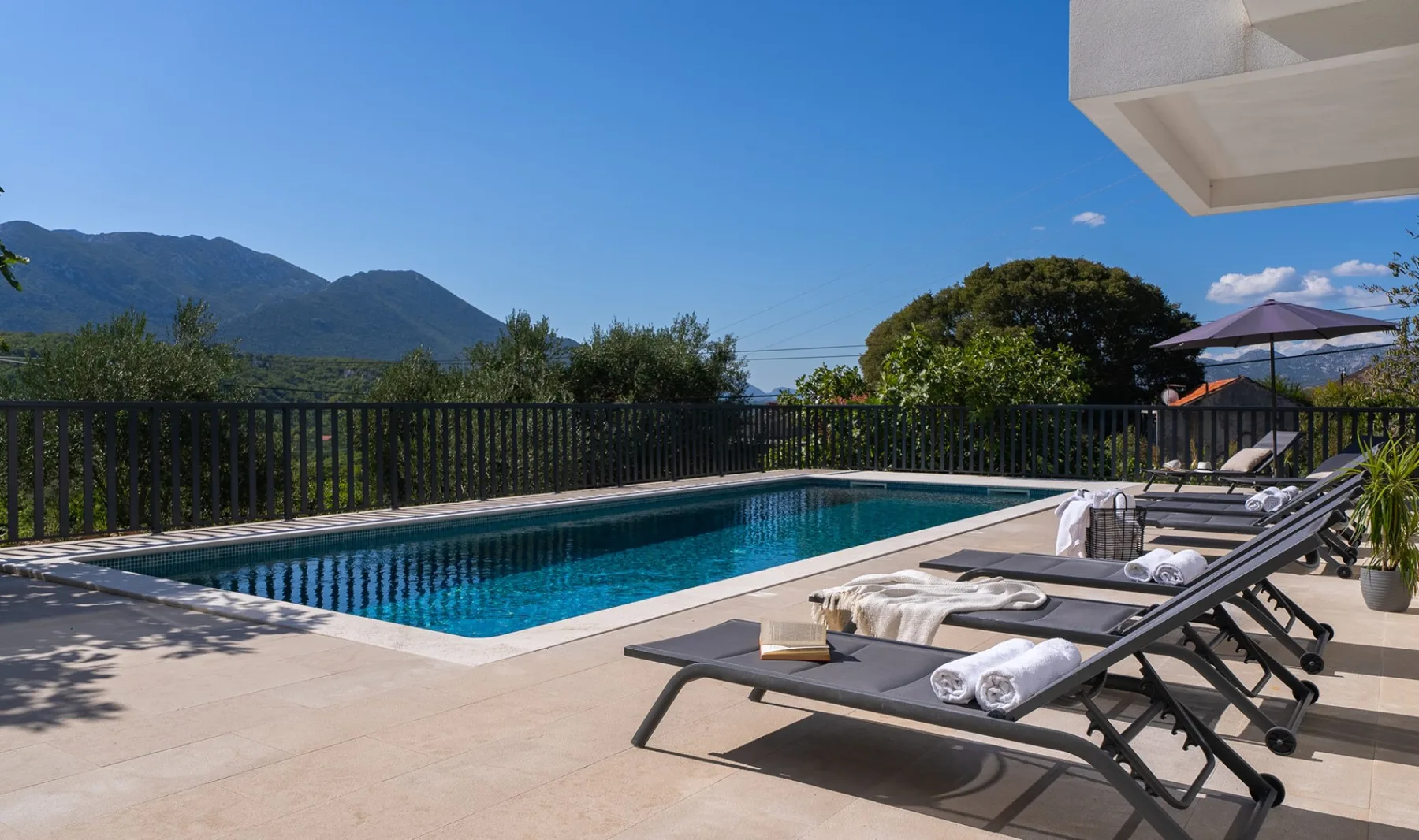 An outdoor swimming pool with lounge seating, greenery, and mountains in the distance.