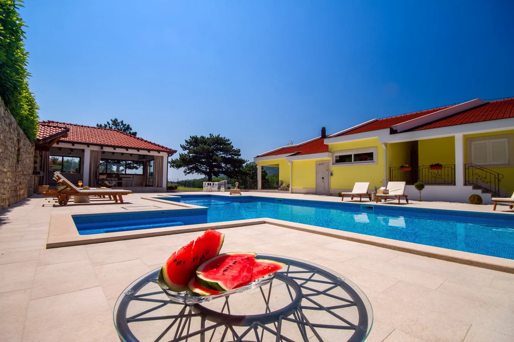 An outdoor swimming pool beside a yellow building and a shaded seating area.