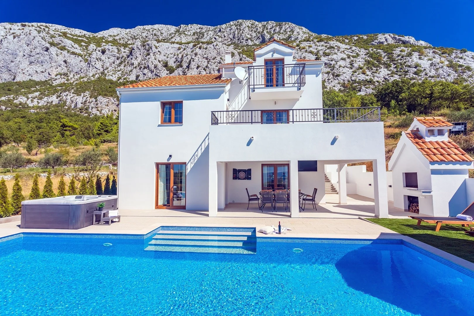 A white two-story house stands behind a swimming pool, with patio seating.
