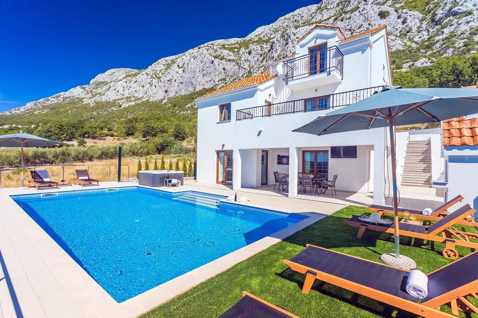 A white villa stands beside a swimming pool, with sun loungers on the patio.