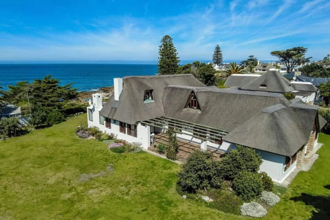 A thatched-roof house sits on a green lawn above the ocean, with trees and neighboring buildings nearby.