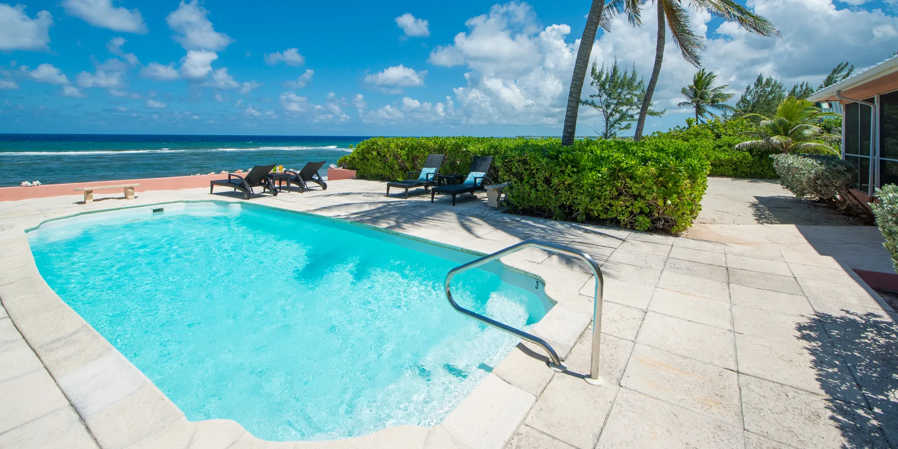 Una piscina junto a un patio pavimentado, con la playa y el océano al fondo.