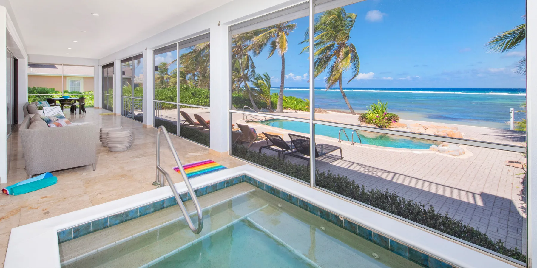 Una habitación cerrada con vidrio da a una piscina, palmeras y el océano.