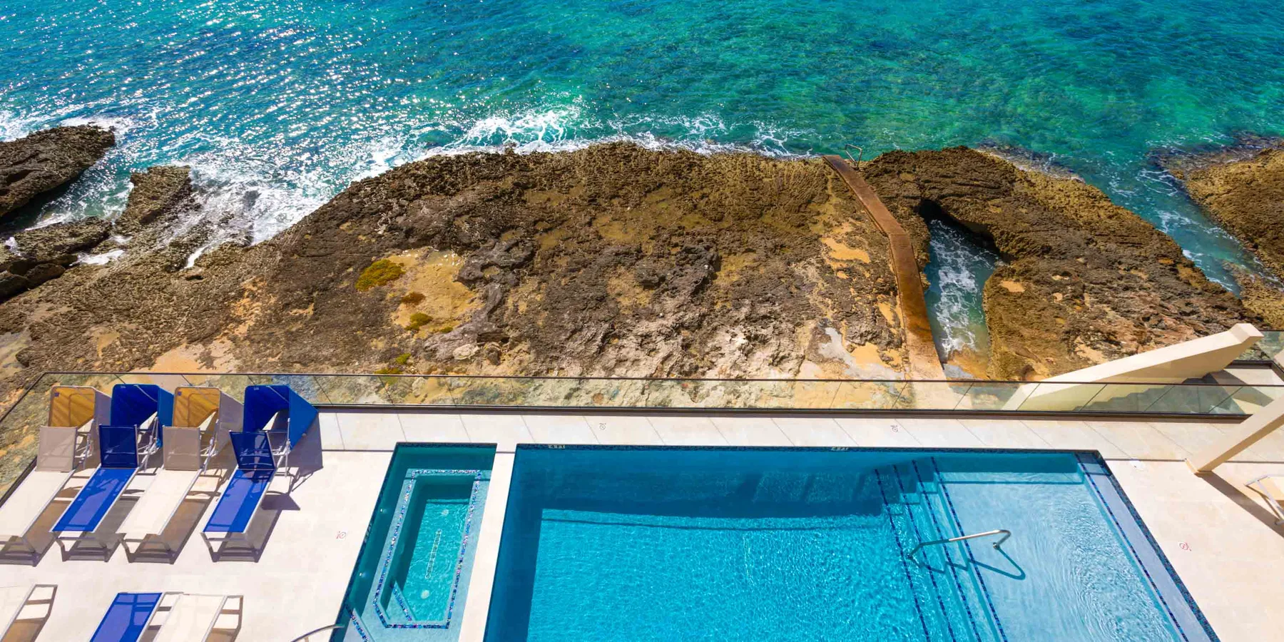 Una piscina y tumbonas dan a una costa rocosa y aguas turquesas.