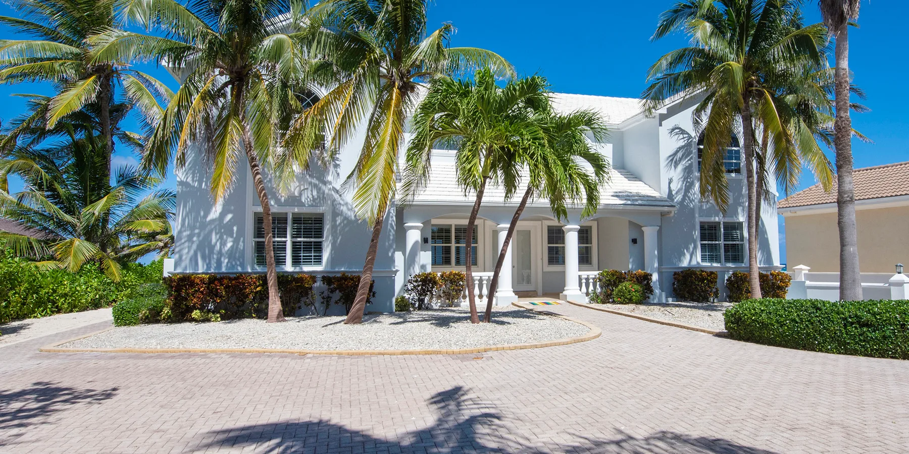 Un edificio blanco de dos plantas se alza detrás de palmeras junto a un patio pavimentado.