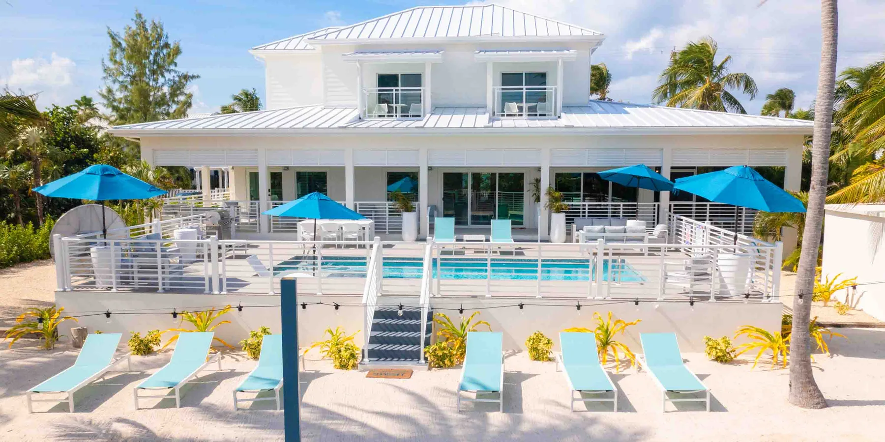 A white house overlooks a swimming pool and lounge chairs on a sunny patio.