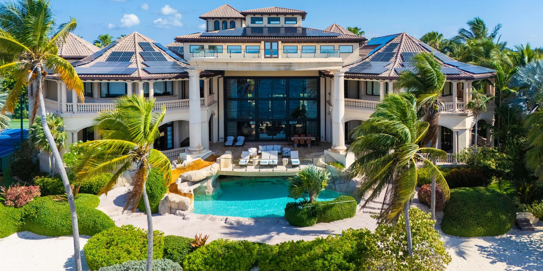 Un gran edificio de estilo turístico da a una piscina rodeada de palmeras y jardines.