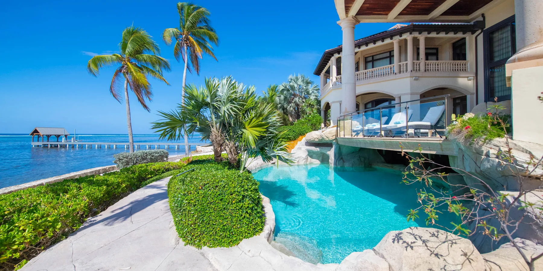 Una villa junto al mar da a una piscina turquesa, con palmeras y un muelle cerca.