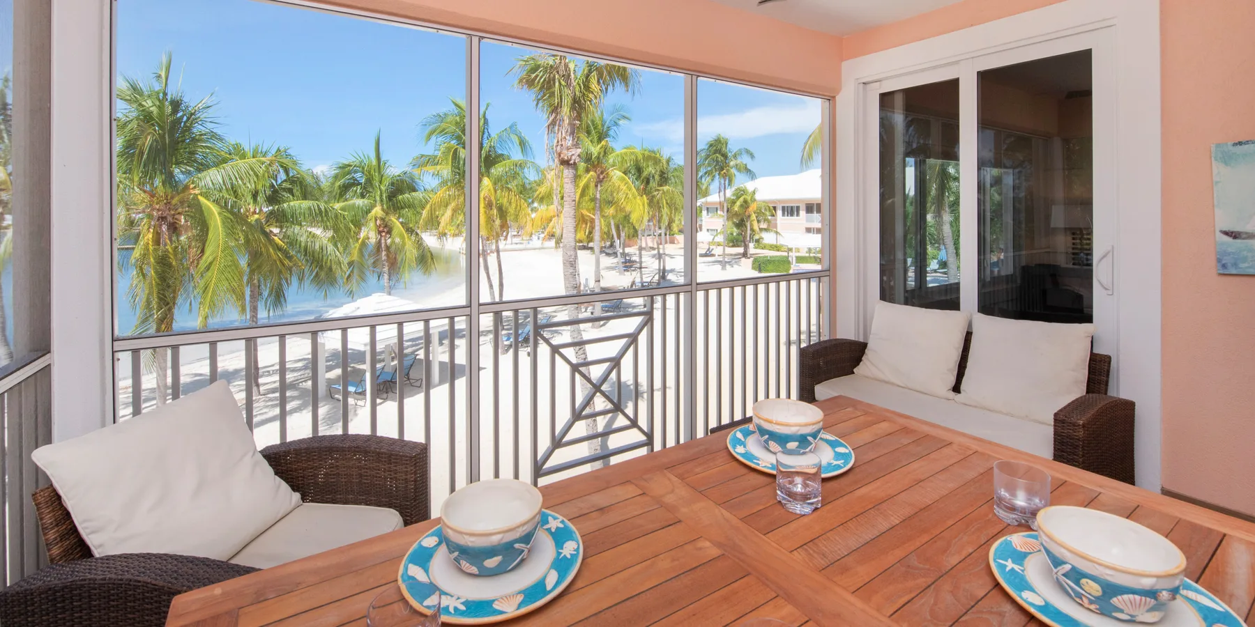 A bright sitting area with a wooden table beside large windows overlooking palm trees.