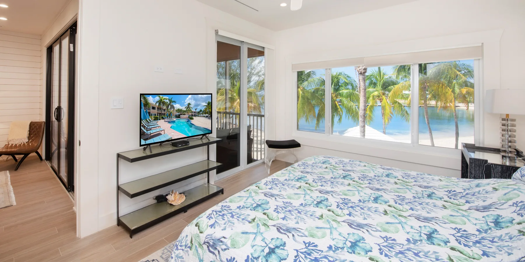 A bright bedroom with a floral bedspread, television, and palm trees visible through the windows.