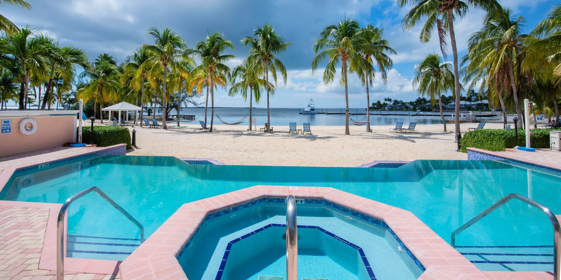 A swimming pool with a small central spa overlooks a palm-lined beach.