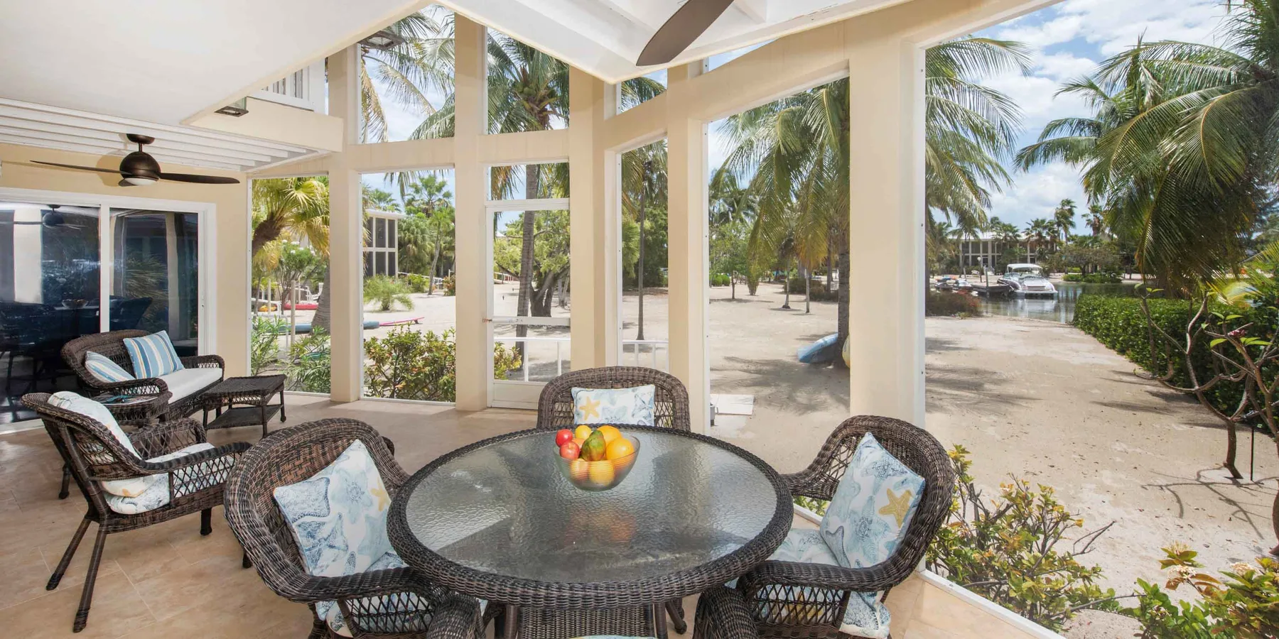 A screened patio with wicker seating overlooks palm trees and a sandy waterfront.