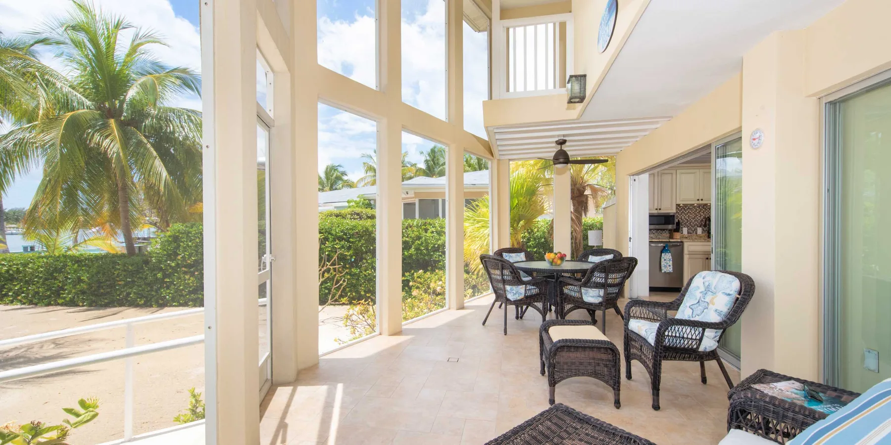 A covered patio with seating and large windows overlooks a palm tree and greenery.