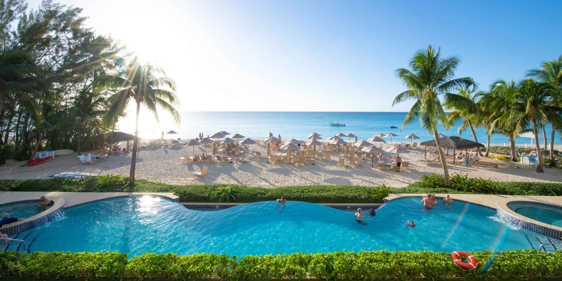 Una piscina frente a la playa da a una orilla arenosa con palmeras y asientos.