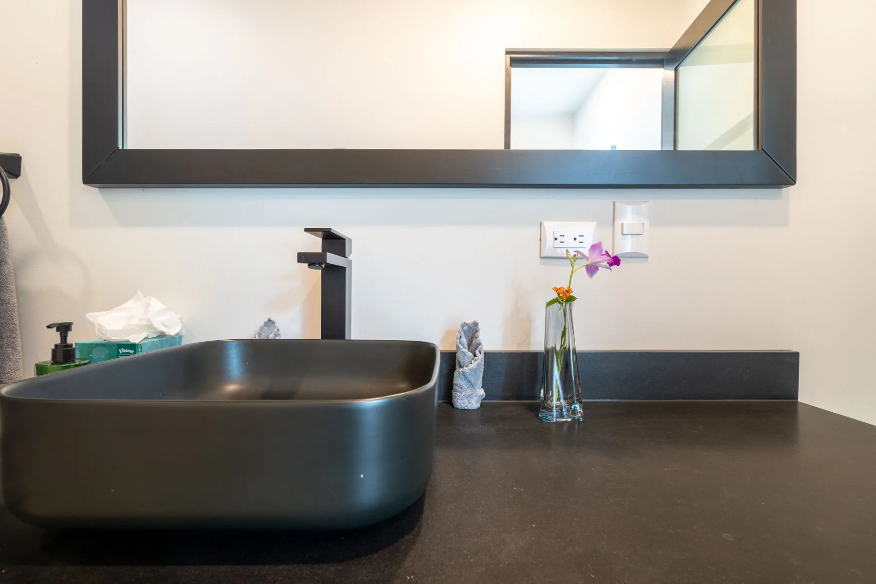 A bathroom vanity with a dark vessel sink, faucet, and flowers.