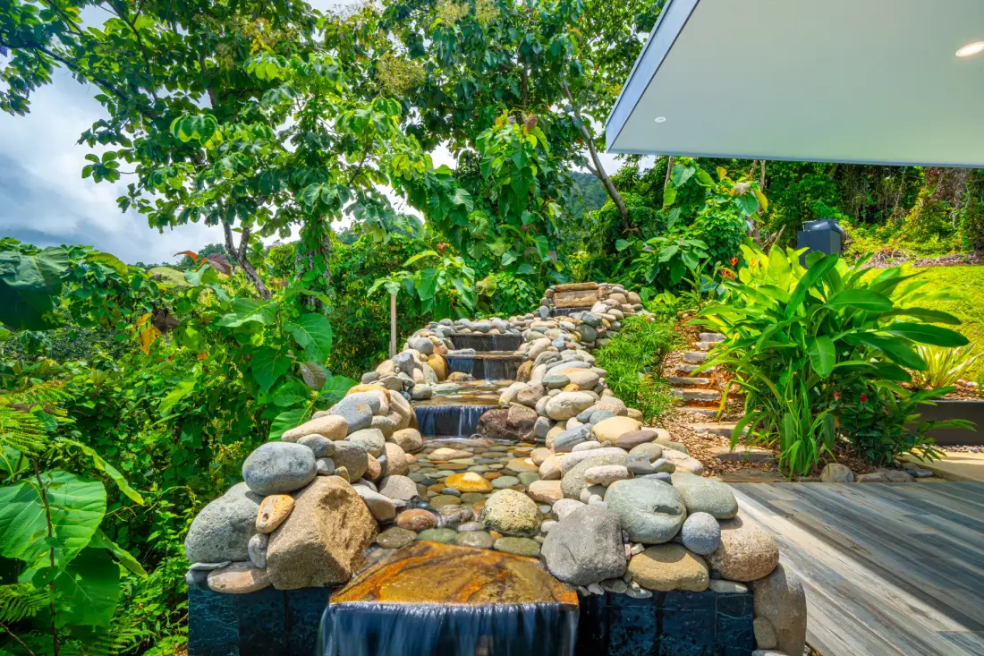 A garden stream cascades over rocks beside a lush tropical walkway.