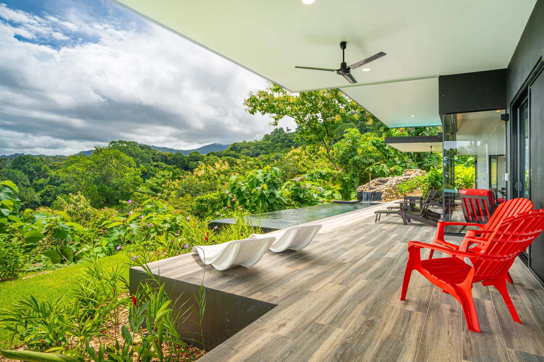 A covered patio with bright seating overlooks a pool and lush green hills.