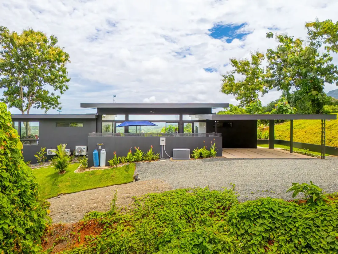 A modern house stands beside a gravel driveway, with trees and greenery around it.
