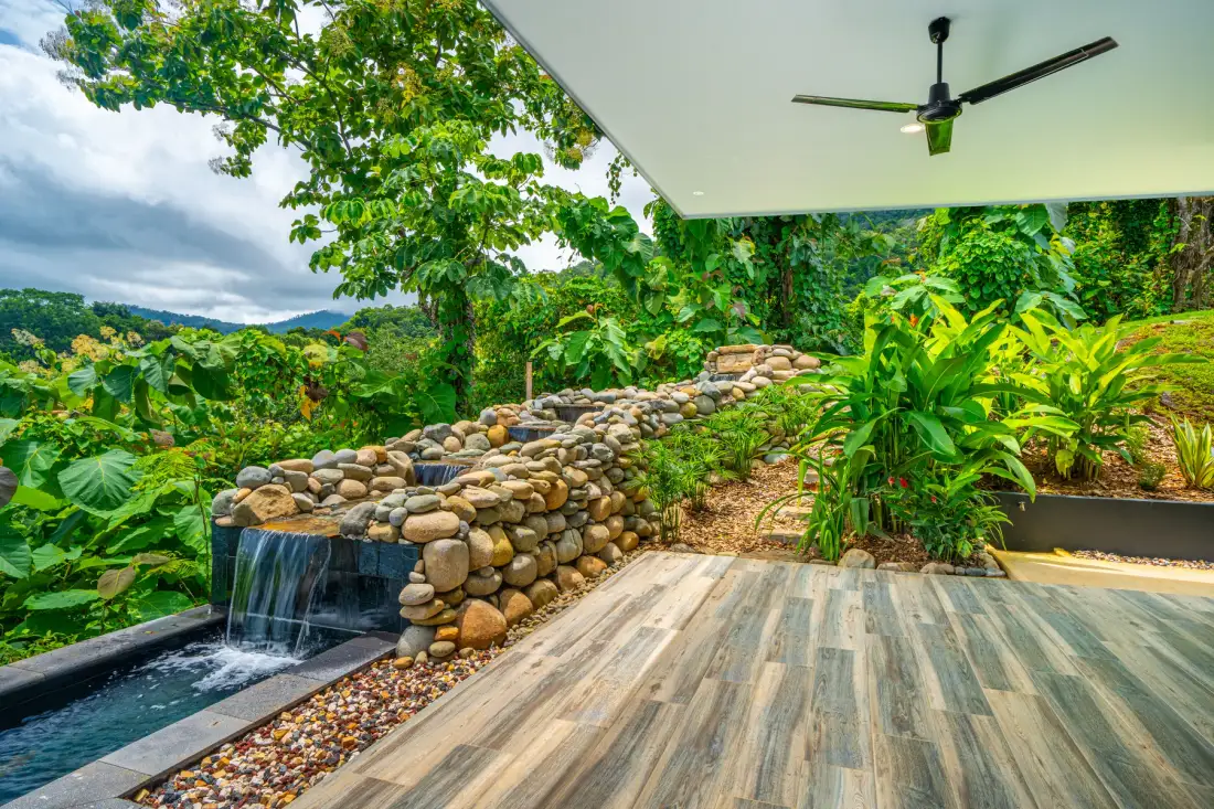 A covered outdoor patio overlooks a stone water feature amid lush greenery.