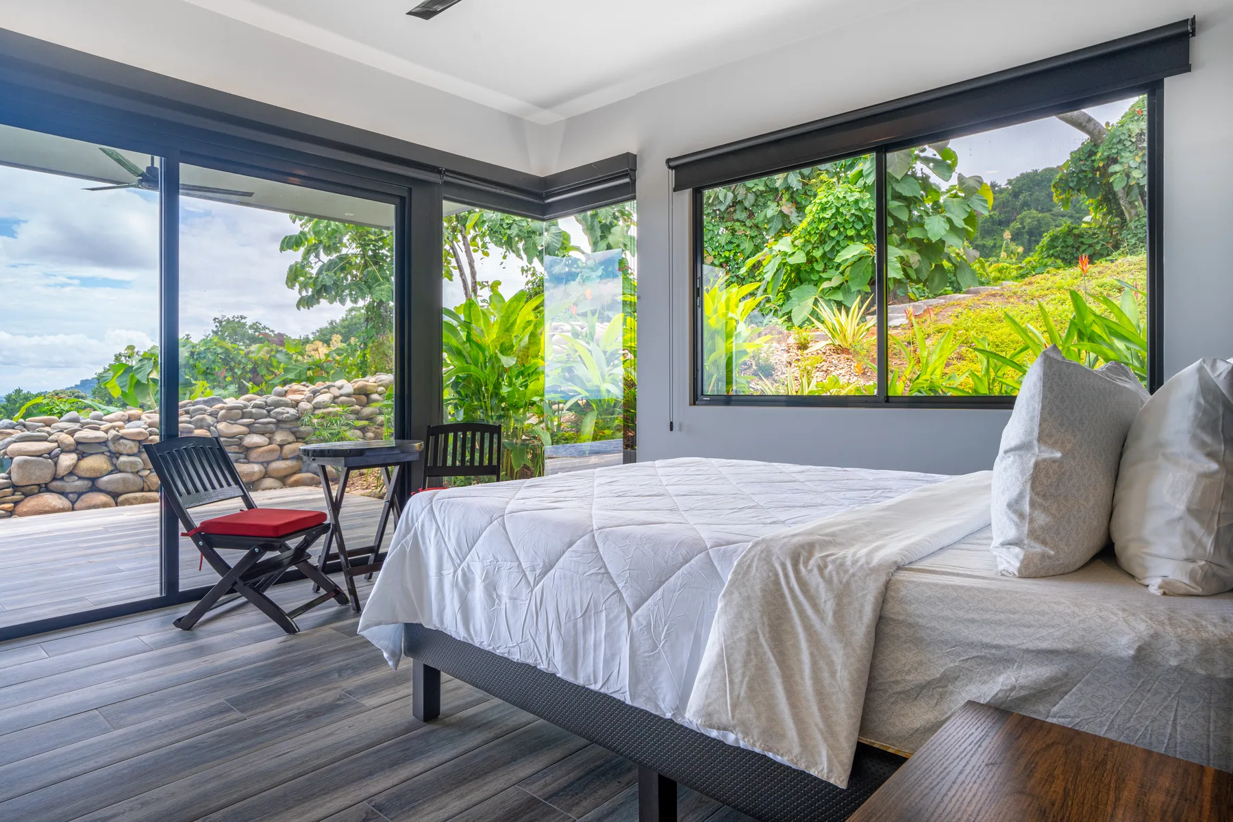 A bedroom with a bed beside large windows overlooking lush greenery.