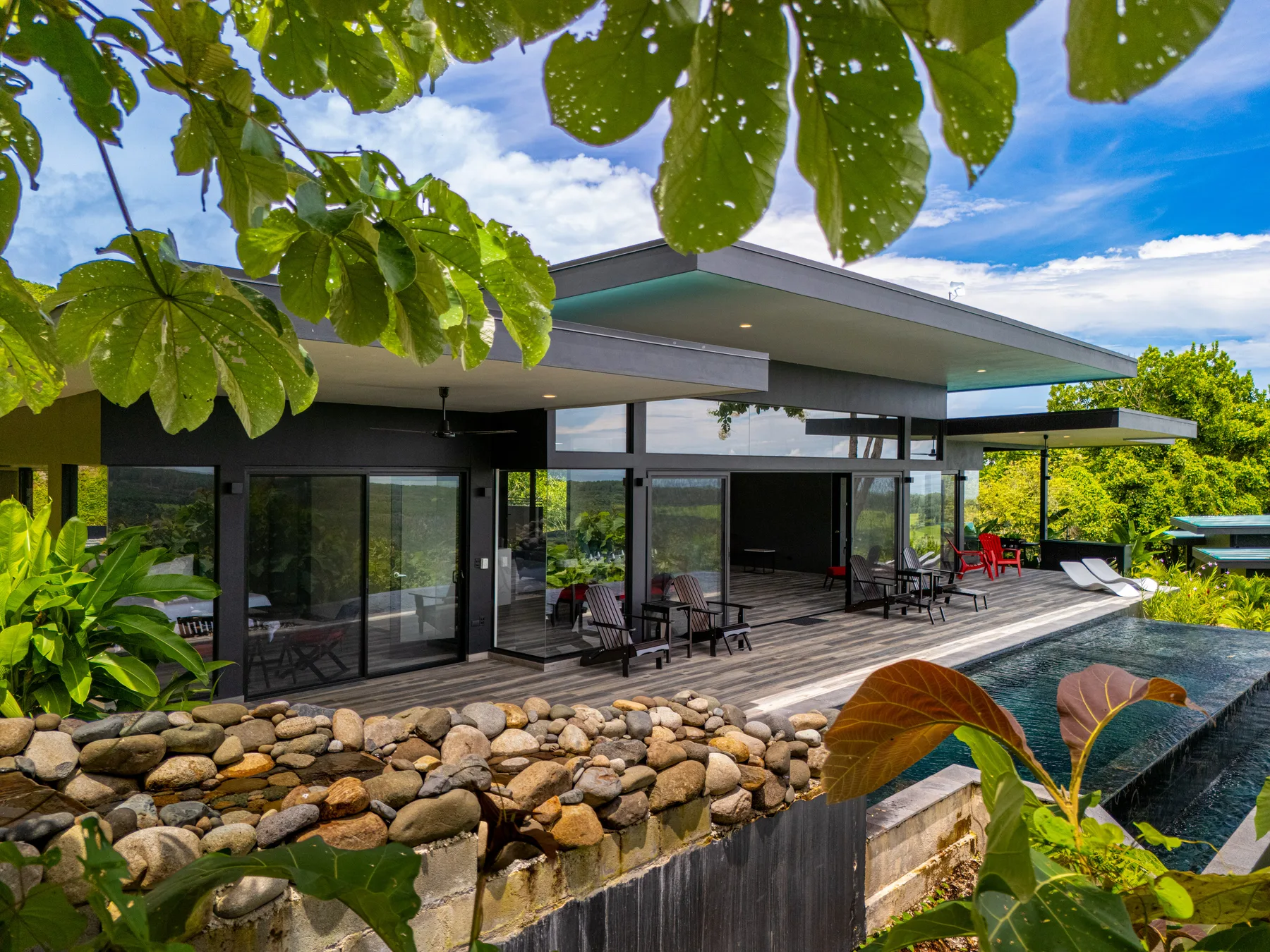 A modern house opens onto a wooden deck beside a swimming pool and landscaped garden.