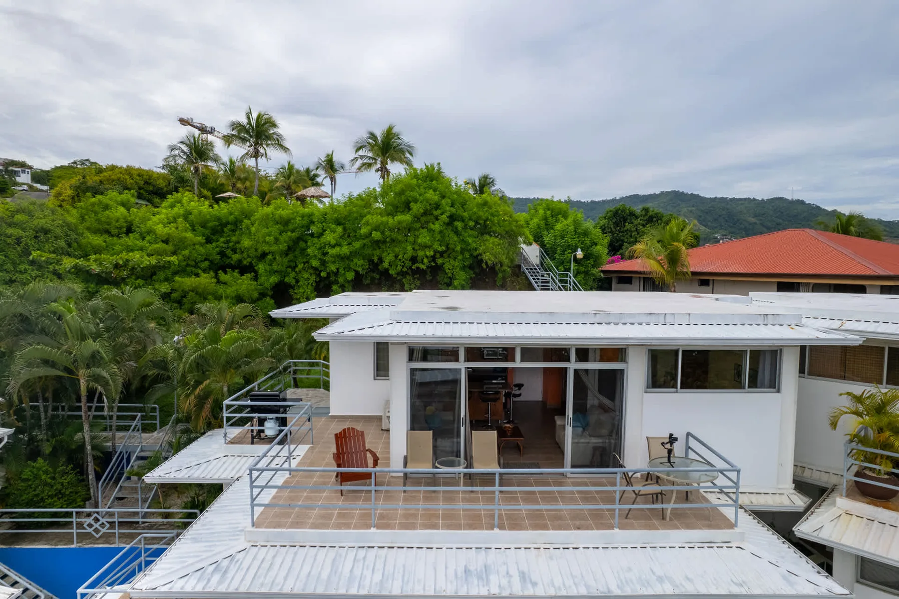 A modern house opens onto a balcony with glass railings, surrounded by tropical greenery.