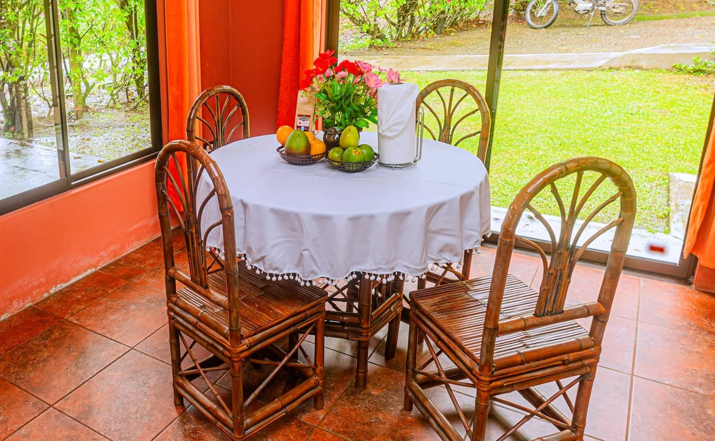 A dining table with wicker seating sits beside large windows overlooking greenery.