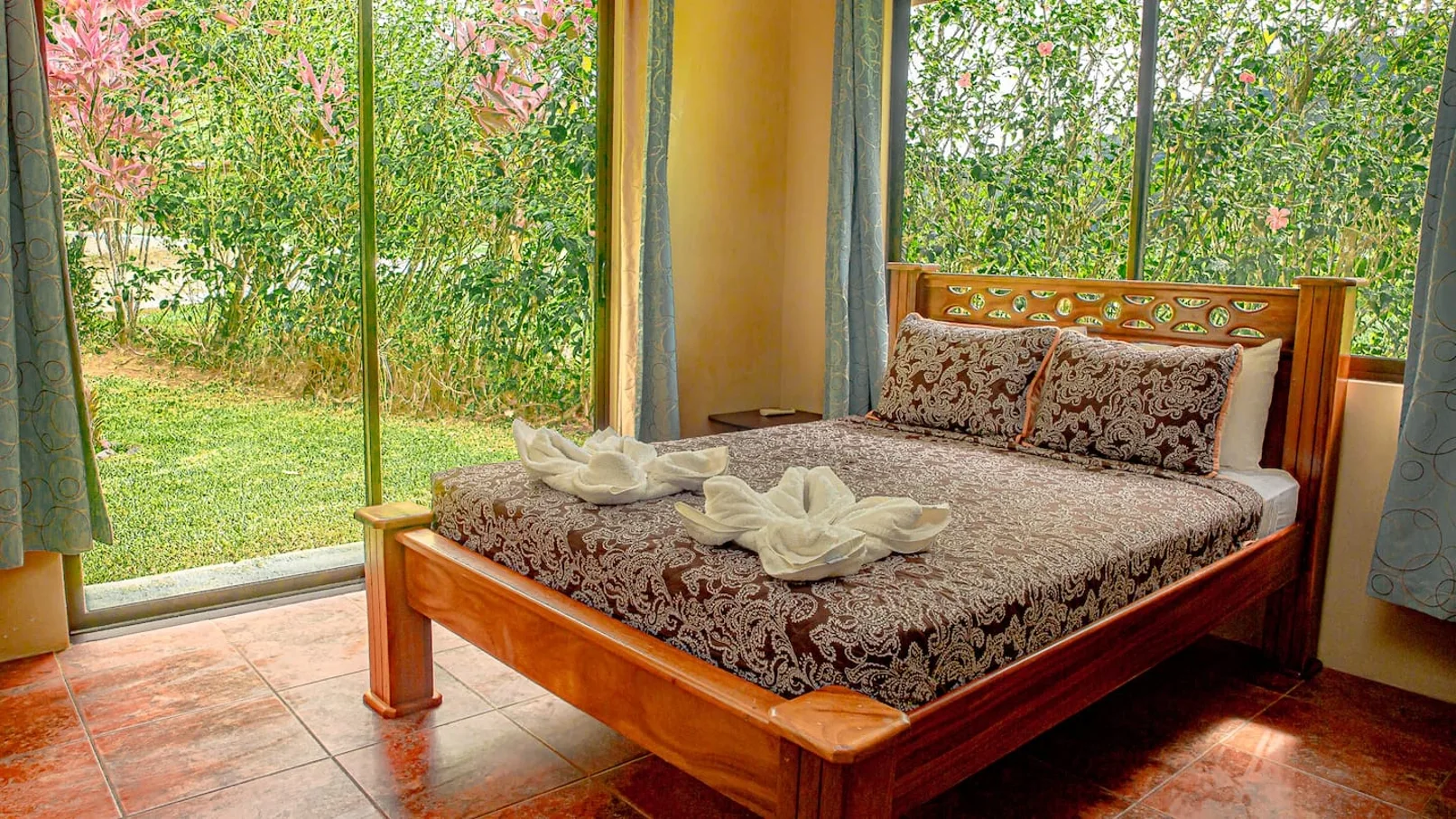 A bedroom with a wooden bed beside large windows overlooking greenery.